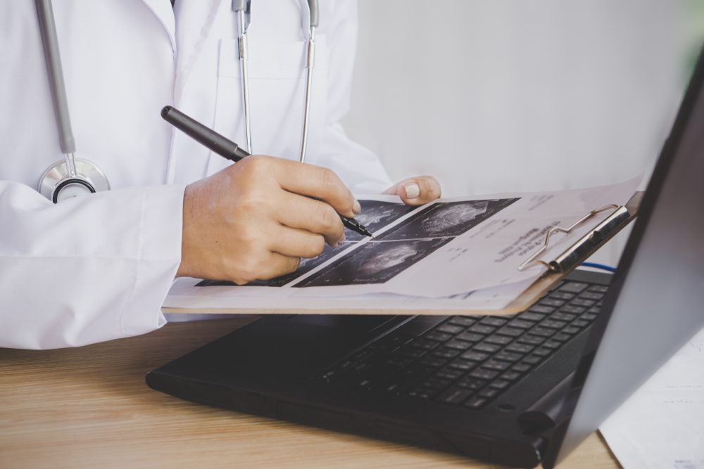 doctor holding pen in hand analyzing x-ray medical picture while working on computer laptop on desk 