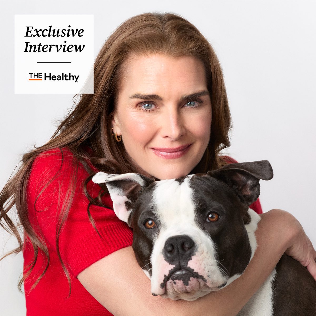 A woman with long brown hair, wearing a red top, smiles while hugging a black and white dog. In the top left corner, text reads "Exclusive Interview" and "The Healthy.